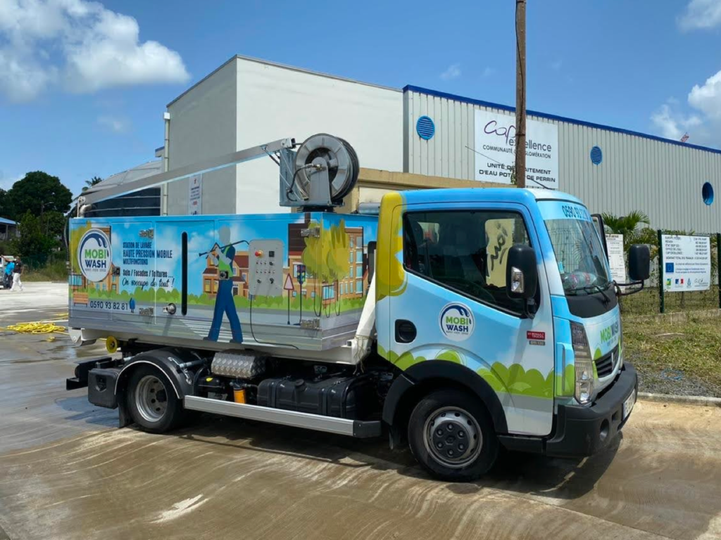 Camion équipé d’un système de lavage haute pression Mirage sur berce ampliroll, avec habillage illustré et bras enrouleur, stationné devant un bâtiment industriel.